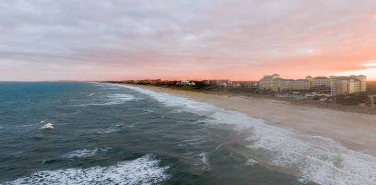 Aerial view of a serene coastal scene where the ocean's foamy waves meet the sandy beach, overlooked by buildings under a pastel-colored sky, Amelia Island, Florida, United States.