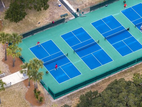 Aerial view of vibrant blue tennis courts contrasting with the lush green surroundings, capturing players in action on a sunny day, Amelia Island, Florida, United States.