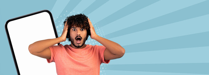 A man stands in front of a plain background. He holds his head with both hands and looks surprised. A large smartphone is beside him, and colorful patterns fill the space around him. © Prostock-studio