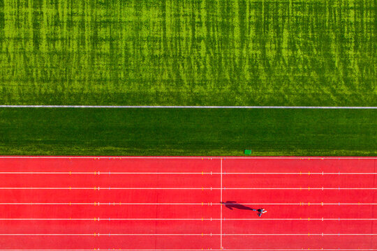 Aerial view of vibrant red running tracks contrast sharply with the adjacent verdant green field, a lone figure casting a shadow on the track, Warsaw, Masovian Voivodeship, Poland.