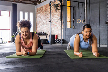 Diverse female workout partners in tanks holding forearm plank on green mats in gym weights visible