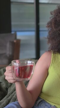 Vertical video: Pulling back, woman on sofa in lime top holding cup with red tea, talking offscreen