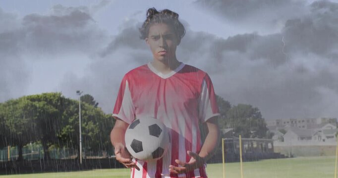 Standing male soccer player handling ball on pitch in red-white kit, storm overlay, yellow poles