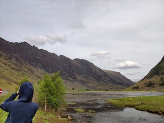 Fototapeta premium Person taking photo of mountain valley landscape with river and cloudy sky in Scotland