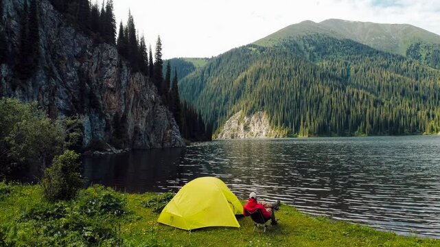 Cinematic low drone flight over camping tent with man sitting on chair against majestic rocky mountains