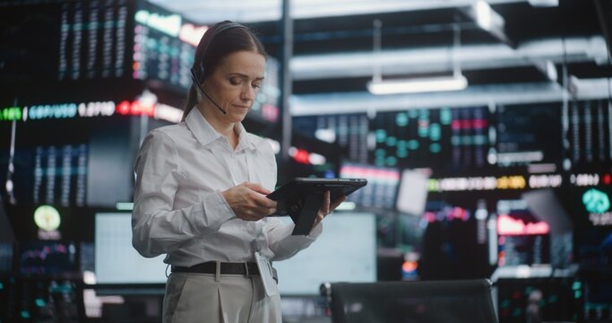 Professional Female Stock Exchange Trader on Trading Floor With Tablet, Monitoring Live Market Data on Digital Screens. Wearing Headset, Analyzes Stock Fluctuations and Coordinates Trading Operations.