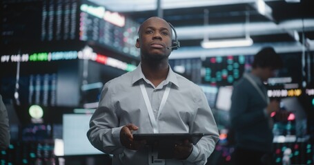 Fototapeta premium Experienced Black Financial Analyst Stands in Modern Trading Center, Carefully Examining Live Global Market Indices on Tablet While Connected Through Professional Headset. Expert Decision Making.