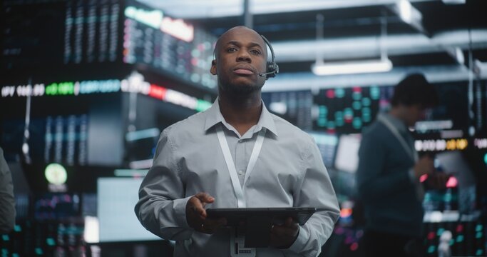Experienced Black Financial Analyst Stands in Modern Trading Center, Carefully Examining Live Global Market Indices on Tablet While Connected Through Professional Headset. Expert Decision Making.