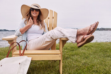 Hat, relax and woman on chair by lake in nature for break on holiday, getaway or weekend trip. Smile, sunglasses and person by river with positive attitude and calm outdoor on vacation in summer.