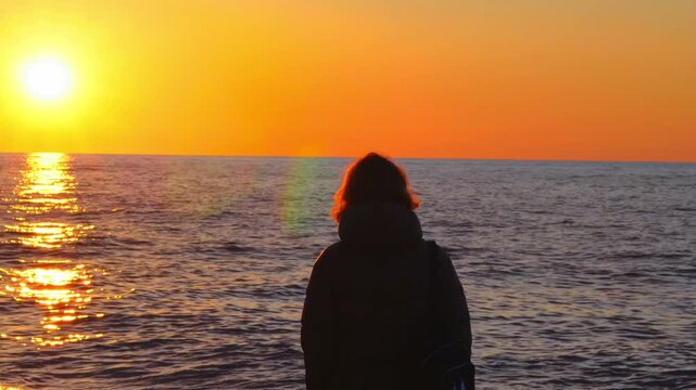 Woman gazes at sunset over ocean, reflections on water, serene atmosphere