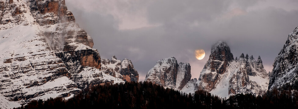 Snow-covered mountain peaks under a cloudy sky, with the moon visible between two peaks. Adamello Brenta Natural Park, Madonna di Campiglio,Trentino,Italy