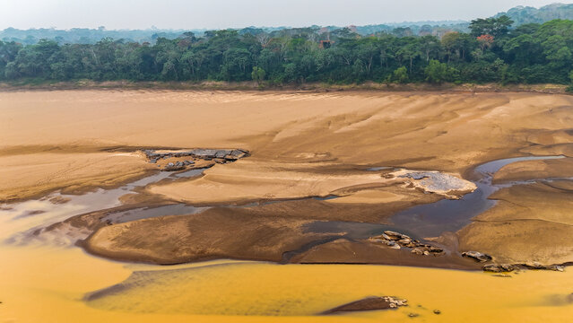 Aerial view of the river's muddy waters weaving through the sandy terrain, contrasting with the lush green rainforest canopy, Puerto Maldonado, Madre de Dios, Peru.