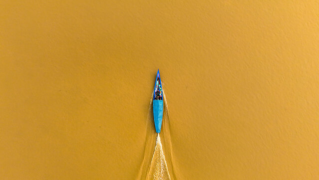 Aerial view of a vibrant blue boat slicing through the golden-brown waters of a river under the warm sun, Puerto Maldonado, Madre de Dios, Peru.