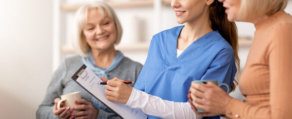 A caregiver meets two elderly women in a home setting. The caregiver holds a clipboard and listens...