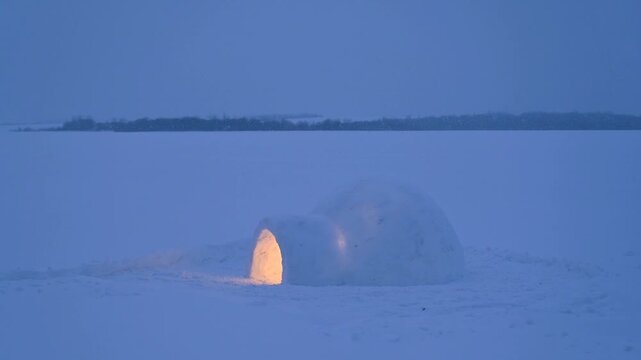 Small igloo standing on frozen snowy landscape during snowfall at blue hour. Winter wilderness scene with falling snow and warm light inside snow shelter