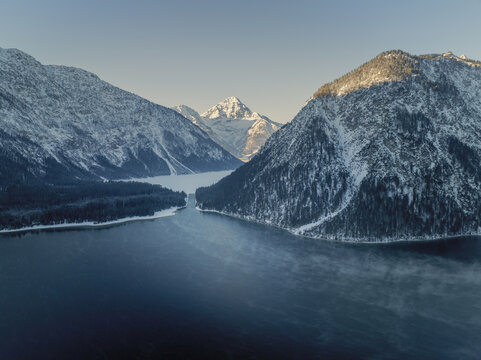 Aerial view of a serene lake nestled between snow-dusted mountains, where the crisp blue waters meet the frosted peaks under a pale winter sky, Tannheim, Austria.
