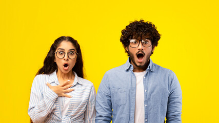 A young couple stands in front of a bright yellow background. They show expressions of shock with wide eyes and mouths open. The woman touches her chest in disbelief. Both wear glasses. © Prostock-studio