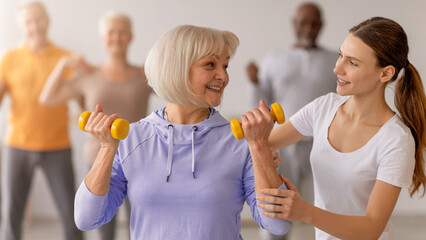 Fototapeta premium A group of older adults participates in a strength training class led by a trainer. One woman lifts yellow weights while the trainer assists her. The atmosphere is supportive and energetic.
