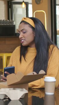Vertical video: Reading blue phone, Indian woman in yellow sweater talking at cafe with food box
