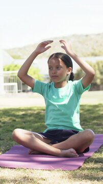 Vertical video: Sitting sunlit girl teal tee raising arms and meditating on purple mat in park