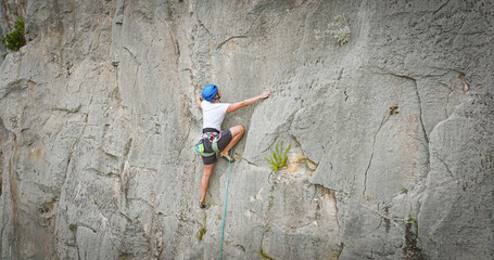 Naklejka premium AERIAL, CLOSE UP: Sport climber stretches for next handhold on a steep limestone wall, wearing a helmet and harness with rope running below. Concentration and agile movement in rugged natural setting.