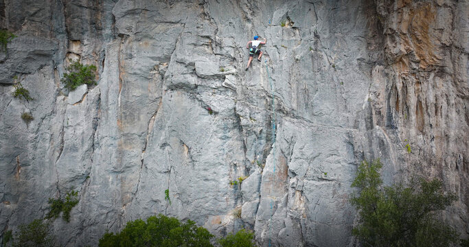 Focused female lead climber moves up a towering limestone face, clipped into a rope for safety. Wide view highlights the scale of a steep rock wall and the exposed, vertical terrain on island of Hvar.