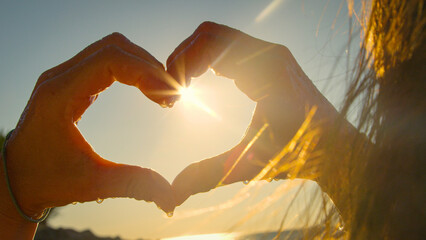 CLOSE UP, LENS FLARE, SILHOUETTE: Unrecognizable woman making a heart shape with her hands,...