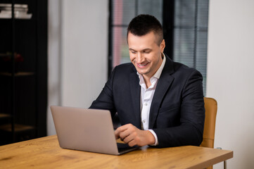 Fototapeta premium Smiling young businessman in suit working on laptop