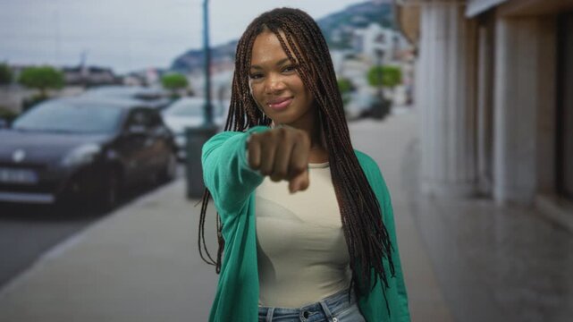 Woman with braided hair wearing green cardigan extends fist toward camera on street lined with parked cars and columns; confidence empowerment.