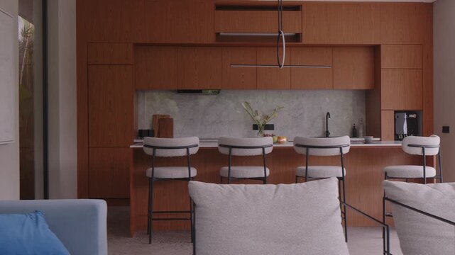 Contemporary kitchen space featuring wooden cabinetry bar stools marble backsplash and open layout with cozy neutral sofa and natural sunlight in living room area.