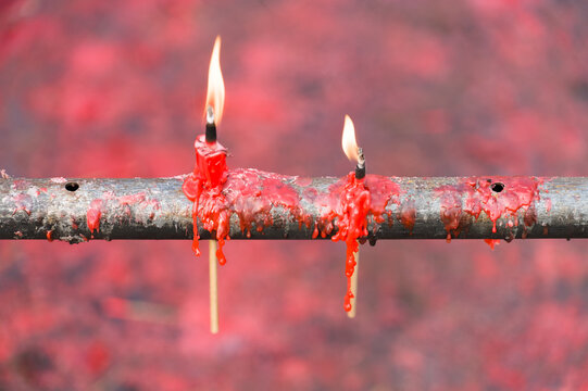 Two vibrant red candles are burning on a weathered metal rod, with melted wax dripping down the sides. The background is a blurred, fiery red, creating a dramatic and intense atmosphere. 
