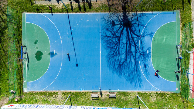 Aerial view of a vibrant blue and green sports court, bisected by the stark shadow of a tree, a geometric spectacle against the green grass, Novi Sad, Vojvodina, Serbia.