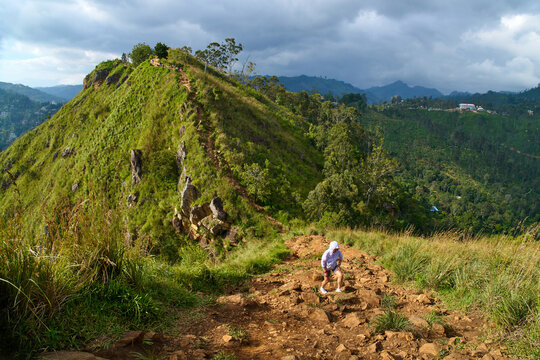 Solo female traveler conquering steep, rugged slope of Little Adam's Peak during golden hour, with majestic Adam's Peak mountain range glowing in warm sunset light, Ella, Sri Lanka.
