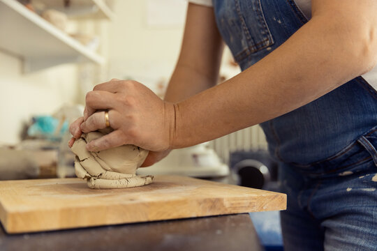 Close-up of a ceramic artist in denim overalls kneading and wedging raw clay on a wooden board in a bright pottery studio. Professional hands preparing clay for throwing on a wheel or handbuilding.