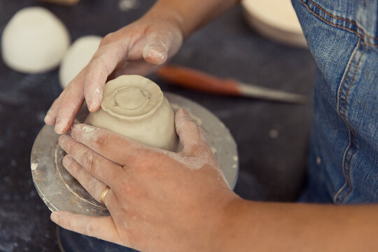 Close-up of a potter's hands carefully shaping a clay bowl on a spinning pottery wheel in a rustic studio. The artisan wears denim overalls, highlighting the authentic craft and handmade process.