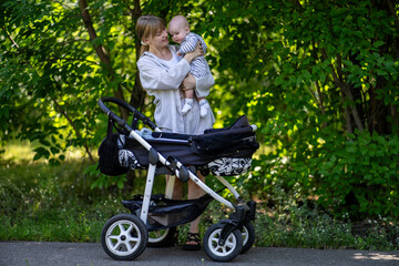 Fototapeta premium A young mother walks in a summer park with a baby in her arms and a stroller