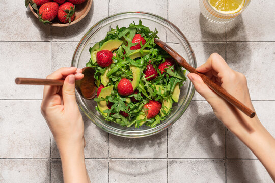 Female hands mixing fresh summer salad with avocado arugula and strawberries in glass bowl on gray tile table, top view healthy homemade meal concept