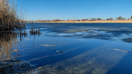 Early spring landscape with a frozen river or lake, clear blue sky reflected in the melt water, and dry reeds on the shore. Ukraine, Dnipro