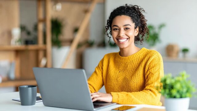 Happy young Black woman working on a laptop in a bright modern home office setting while smiling at the camera