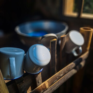 Metal cups and simple kitchen utensils arranged on a wooden rack inside a rustic village kitchen. Soft natural lighting highlights the authentic atmosphere of traditional rural living.
