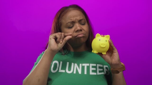 African american volunteer woman holds yellow piggy bank and pinches fingers in purple studio; thrift caution.
