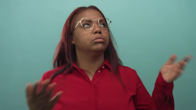 Woman lifts both hands to fan her face in a studio setting while wearing glasses and a red shirt; discomfort.