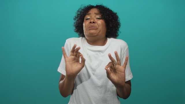 Young african american woman wearing white t shirt cringing while holding ok sign in a teal studio; disgust.