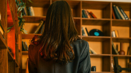 Woman with long brown hair in a suit jacket stands in front of a wooden bookshelf © shine.graphics
