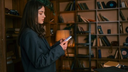 Young woman in a suit using her smartphone in a cozy library with bookshelves © shine.graphics