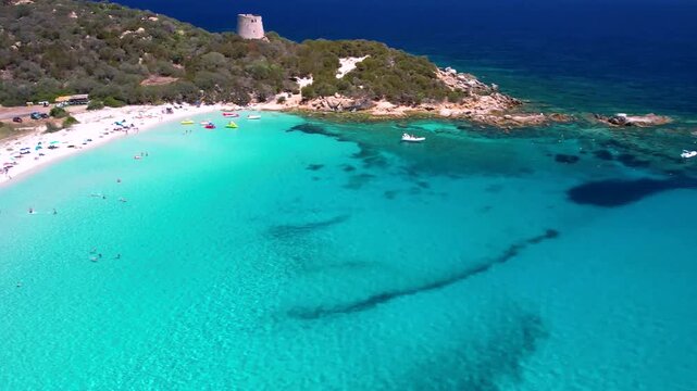 White sailing yacht in turquoise sea aerial orbit shot with Saracen tower view at Cala Pira beach Sardinia European tropics luxury travel destination 4k cinematic drone footage Italy coast