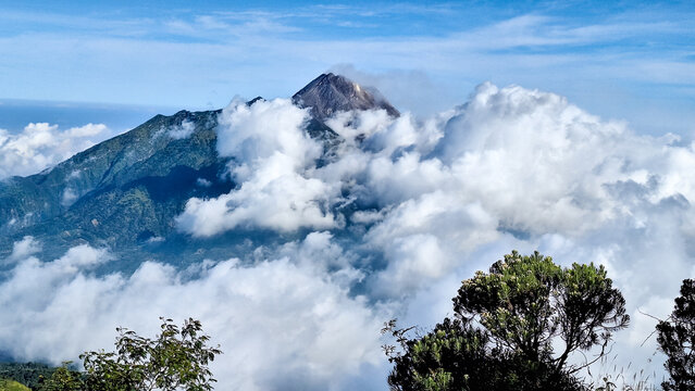 Beautiful landscape of Mount Merapi rising above thick clouds with lush tropical vegetation in Indonesia. The dramatic volcanic mountain scenery shows natural beauty and adventure destinations.