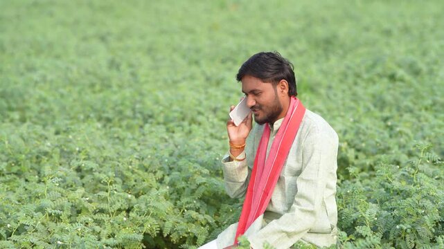 young indian farmer using smartphone sitting at green agricultural field
