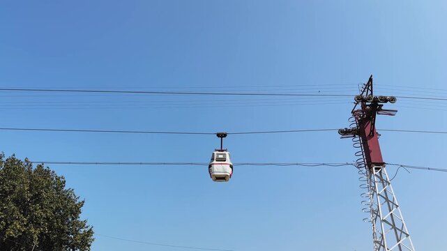 Cable car gondola moving along a ropeway cable against a clear blue sky supported by a metal tower, showing aerial transport used for tourism and travel.