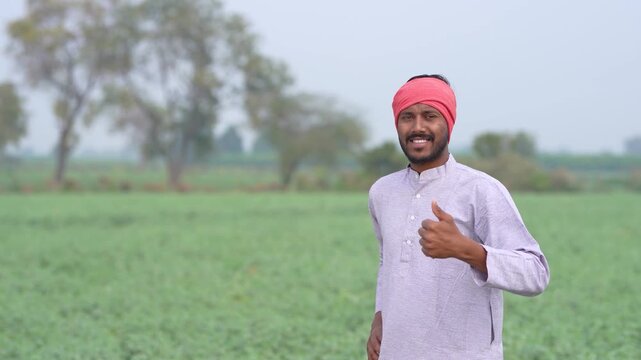 young indian farmer standing and showing thumps up at green agricultural field
